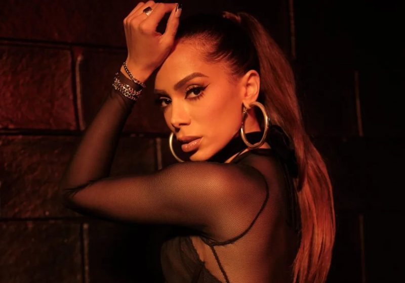 In the image, Anitta is wearing a black blouse with hoop earrings and looking to the left. 