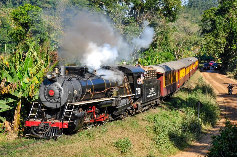 Foto de uma locomotiva maria fumaça puxando dois vagões de um trem de passeio. A locomotiva é preta, com detalhes vermelho e de sua chaminé sai bastante fumaça cinza. Os vagões onde vão os passageiros são de madeira, nas cores vermelha e amarela. A locomotiva passa no meio de uma área de bastante vegetação. Do lado direito, uma estrada de terra, onde circulam alguns carros e uma moto. 