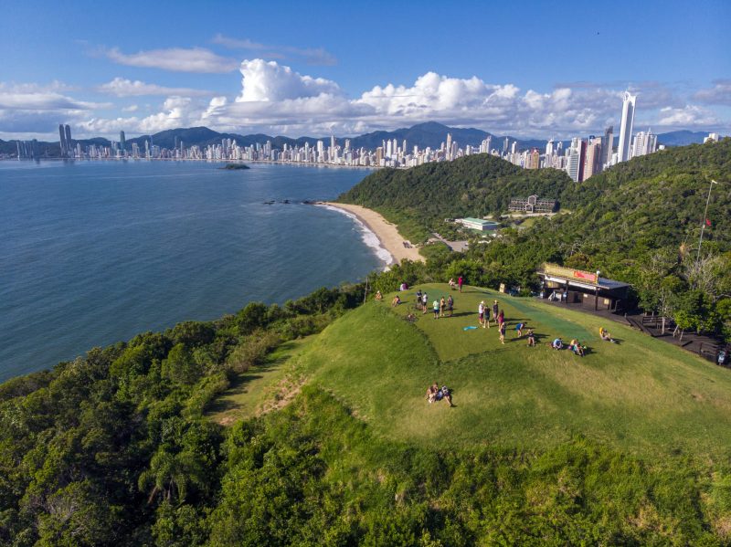 Foto aérea do Morro do Careca, na divisa de Balneário Camboriú e Itajaí. No centro e à direita da foto o morro e muita vegetação. No canto esquerdo e no horizonte, o mar e os prédios da orla central de Balneário Camboriú. Atrás deles, outros morros verdes. O céu está azul. 