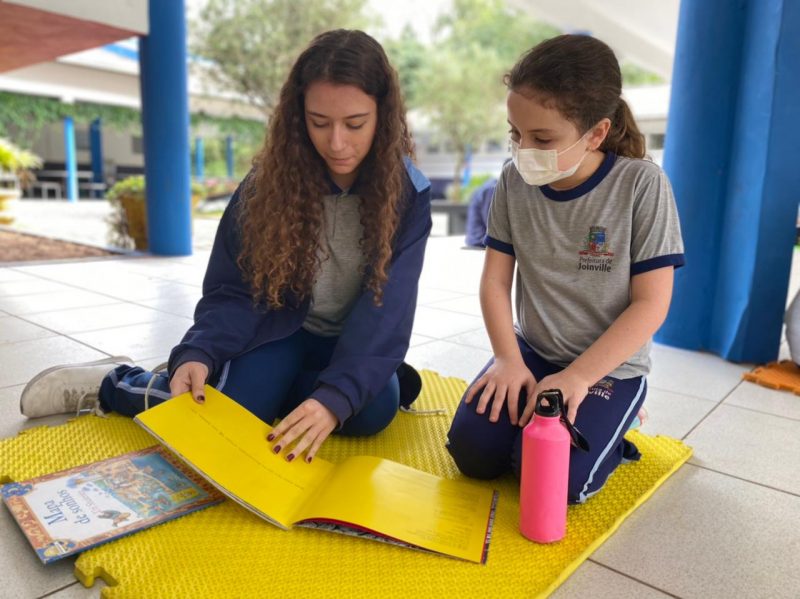 Na imagem, duas alunas fazem a leitura de um livro