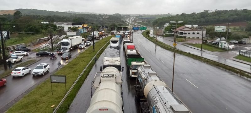 Imagem mostra caminhões parados em uma fila na rodovia. O tempo está nublado