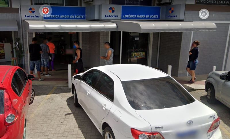 Imagem mostra lotérica Magia da Sorte e algumas pessoas na fila, tem carros estacionados na frente da loteria