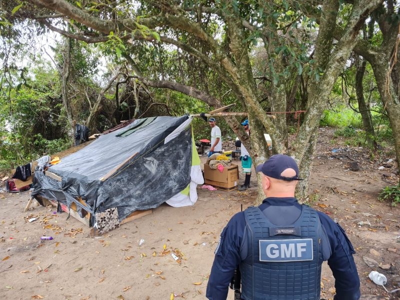 Imagem mostra a barraca coberta de lona e um policial onde é possível ler GMF