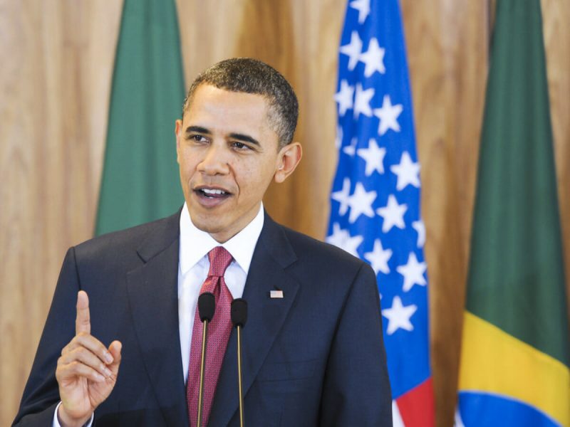 Barack Obama, homem negro, discursando frente a bandeira do Brasil e Eua