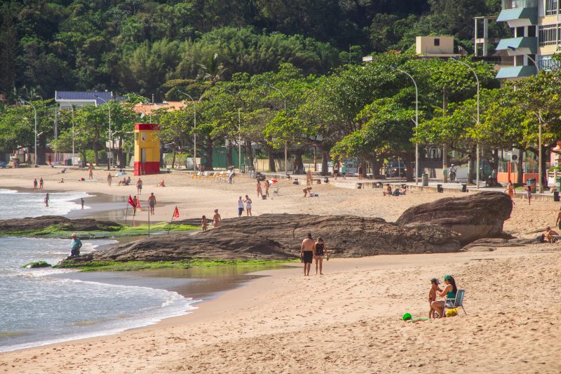 Praia de Cabeçudas, em Itajaí (SC), durante o verão. A foto mostra no canto esquerdo, apenas uma beirada do mar. Centralizado e na maior porção da imagem aparece a faixa de areia, com algumas pessoas, uma rocha que pega uma trecho de areia e mar e ao redor árvores. 