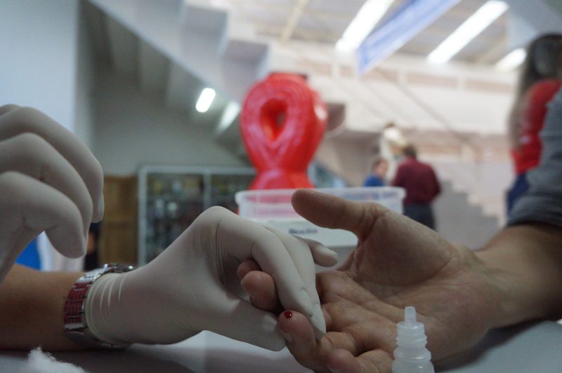 Imagem mostra três mãos e uma delas segura a outra para fazer teste de sangue