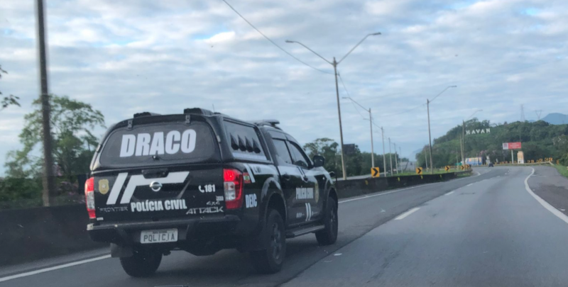The image shows a black civilian police car with Draco on the highway visible on the rear window.  the sky appears with many clouds