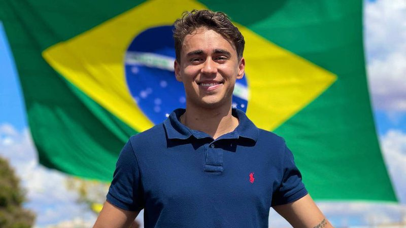 Na imagem, Nikolas Ferreira aparece sorrindo na frente da bandeira do Brasil