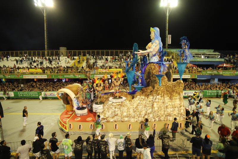 Desfile de carro alegórico na Passarela Nego Quirido no Carnaval. O carro mostra uma sereia de cabelo azul em cima da areia e de conchas de búzios, com participantes fantasiados com dores vivas.