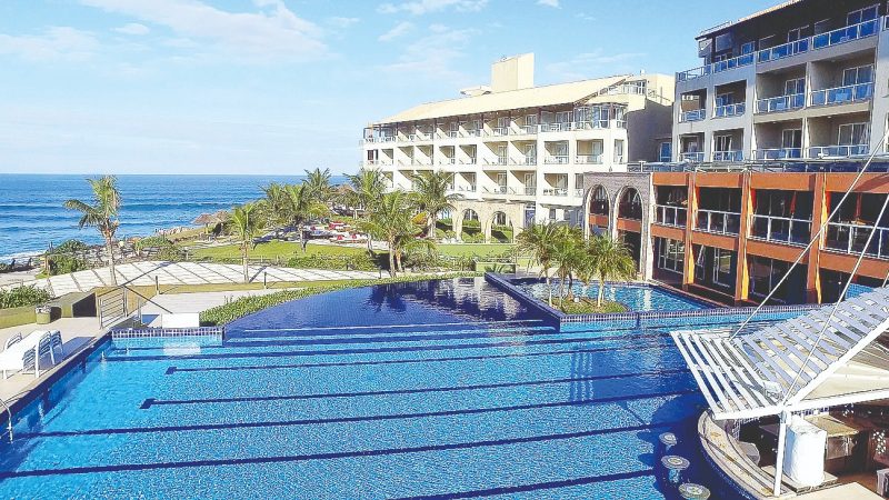 Foto do resort Costão do Santinho, em Florianópolis. Em primeiro plano aparece uma grande piscina azul. Do lado direito e ao fundo, dois prédios do resort. No centro e atrás da piscina, uma área gramada com algumas árvores e mais ao fundo ainda o mar. 