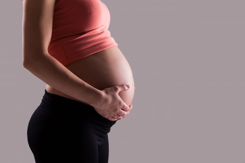 Mulher grávida usando blusa rosa e calça preta segurando a barriga de lado, para ilustrar matéria sobre aborto em grávida errada