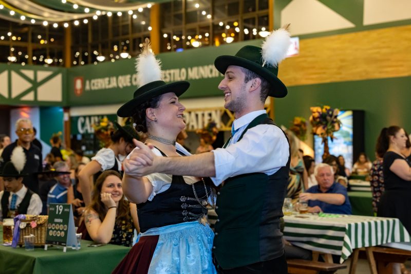 Foto de um casal jovem de pele clara e com trajes típicos germânicos dançando de mãos dadas. Eles estão em um pavilhão fechado do Parque Vila Germânica, em Blumenau. Atrás deles há mesas com pessoas e no teto tecidos coloridos e luzes. 