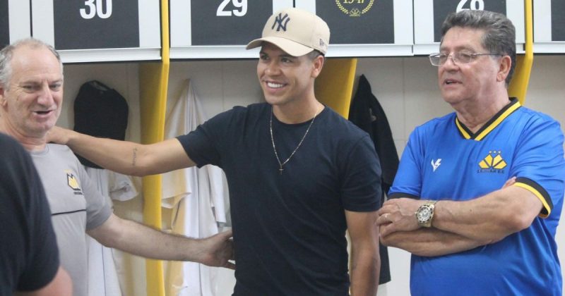 Imagem mostra Marquinhos Gabriel usando uma camiseta preta, boné bege e acompanhado de dois homens no vestiário 