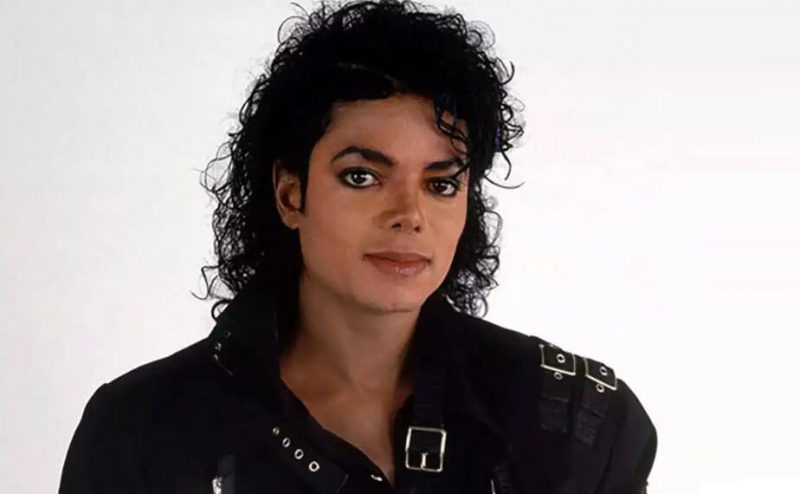 Michael Jackson - homem de pele clara, na altura dos ombros, cabelos pretos ondulados, usa jaqueta de couro preta com fivelas.