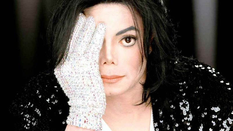 Cantor Michael Jackson - homem branco de cabelos curtos e pretos cobrindo metade do rosto com uma luva branca cheia de brilhos. Imagem é utilizada para ilustrar matéria sobre necrópsia de Michael Jackson