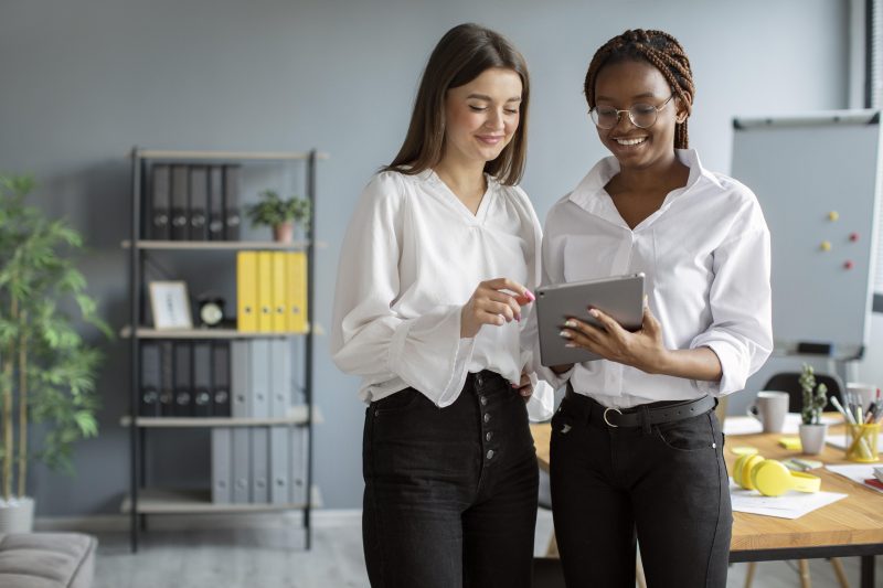 Foto mostra duas mulheres vestindo roupa social olhando para um tablet. Imagem ilustra matéria sobre desigualdade salarial entre homens e mulheres.