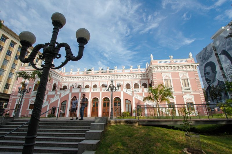 Foto da fachada do Palácio Cruz e Sousa, em Florianópolis. O prédio é rosado, com as portas arredondadas e de madeira. O edifício é bem ornamentado com detalhes em branco. À frente dele, uma escadaria cinza. Na lateral direita um mural em preto e branco com a pintura de Cruz e Sousa, homem negro, poeta que dá nome ao prédio. 