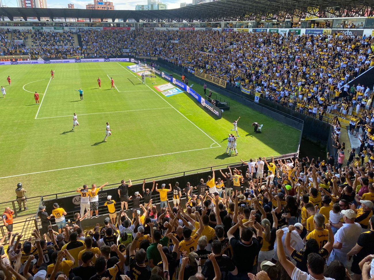 Foto de parte da arquibancada do Estádio Heriberto Hülse, em Criciúma. No meio da foto, aparece parte do campo de futebol com jogadores correndo.