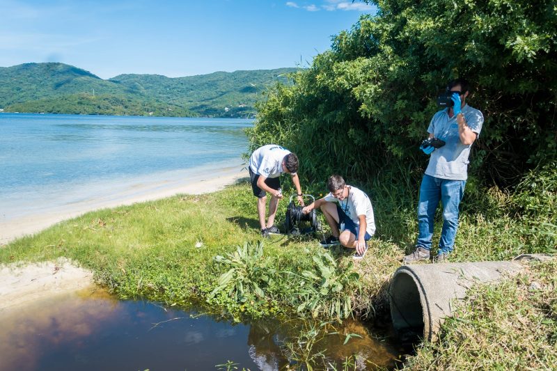 Foto mostra três pessoas em frente a um duto que despeja esgoto em uma lagoa