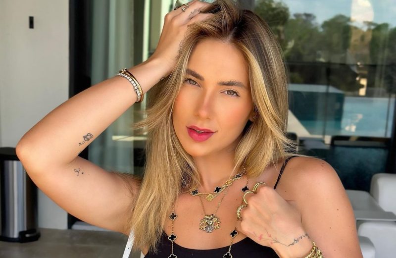 Mulher loira posando com a mão na cabeça. Ela usa um colar com símbolos pretos e bordas douradas, duas pulseiras no braço direito, 3 anéis nos dedos da mão esquerda e uma blusa preta de alcinha