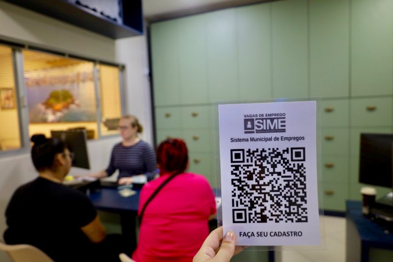 Foto de uma mão segurando um QR code do SIME, enquanto duas mulheres, ao fundo, conversam com funcionária da entidade em avaliação para emprego