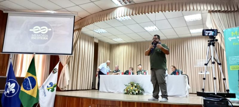 Imagem mostra um homem vestido de verde no canto direito e no canto esquerdo uma tela onde é possível ler Memória Escoteira