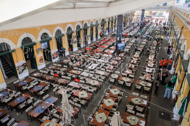 Foto do pátio interno do Mercado Público de Florianópolis, na perspectiva de cima. Aparecem dezenas de mesas de madeira com cadeiras no centro da foto. Nas laterais, aparecem as entradas de cada boxe comercial. A parede é pintada de amarelo, com arco branco em cima. 