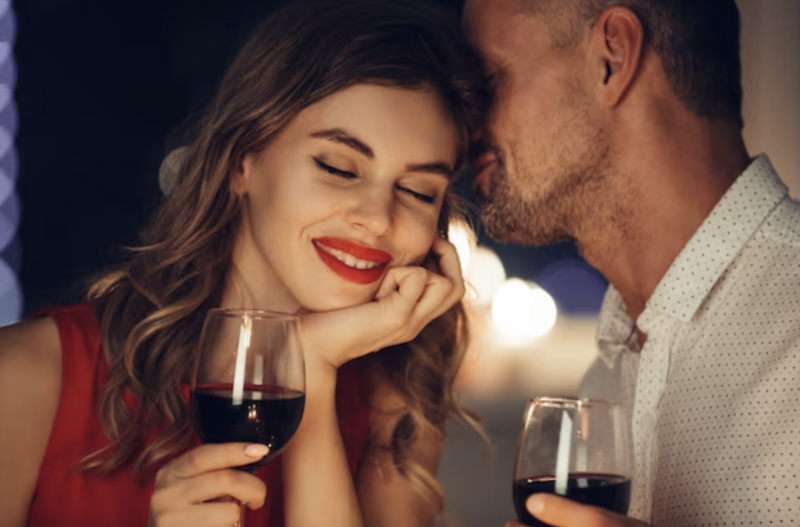 casal com taças de vinhos, ela com os olhos fechados e sorrindo e encostando a cabeça de lado no rosto dele enquanto ele sorri com o olho fechado também 