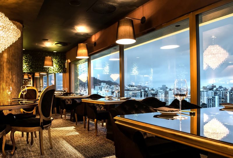 Foto mostra restaurante com mesas próximas a uma grande parede de vidro com vista para a beira-mar de Florianópolis. 