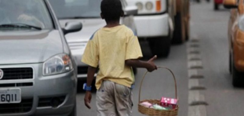 trabalho infantil, foto mostra menino de costas carregando cesta de vine entre carros