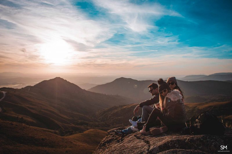 Foto de três pessoas agasalhadas em cima do mirante dos Campos do Quiriri, em Campo Alegre. Ao fundo, morros, céu azul e o pôr do sol. 