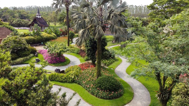 Foto de um bonito jardim com flores e árvores. Ao fundo há uma pequena capela de madeira. 