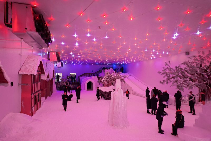 Foto de um espaço fechado com neve, casinhas de esquimó, escorregador e pessoas bem agasalhadas.