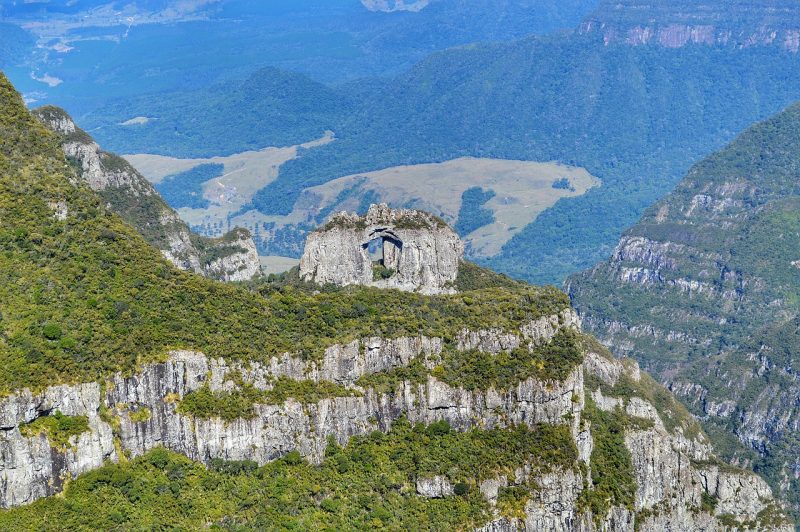 No centro da foto, a Pedra Furada, na Serra Catarinense. Ela é uma rocha alta e larga e possui um vão no meio. Ela está em cima de um morro coberto pela vegetação. Ao fundo, aparecem outros morros. 