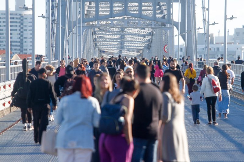 Foto de dezenas de pessoas caminhando pela Ponte Hercílio Luz. Aparece apenas parte da estrutura metálica da ponte e pelas laterais alguns prédios. 