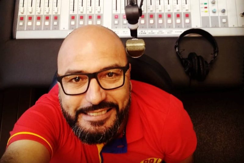 Sérgio Roberto Mattos aparece de camiseta vermelha. Ao fundo, mesa de operação de rádio