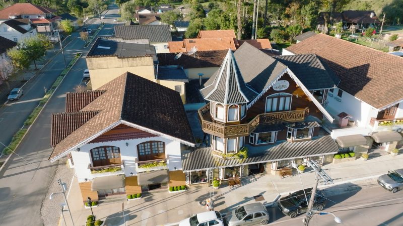 Foto aérea de uma construção da área urbana de Campo Alegre: uma casa em estilo alpino, onde funciona uma loja de artesanato. À frente dela e no canto esquerdo aparecem ruas com alguns carros estacionados. Ao fundo há algumas árvores. 