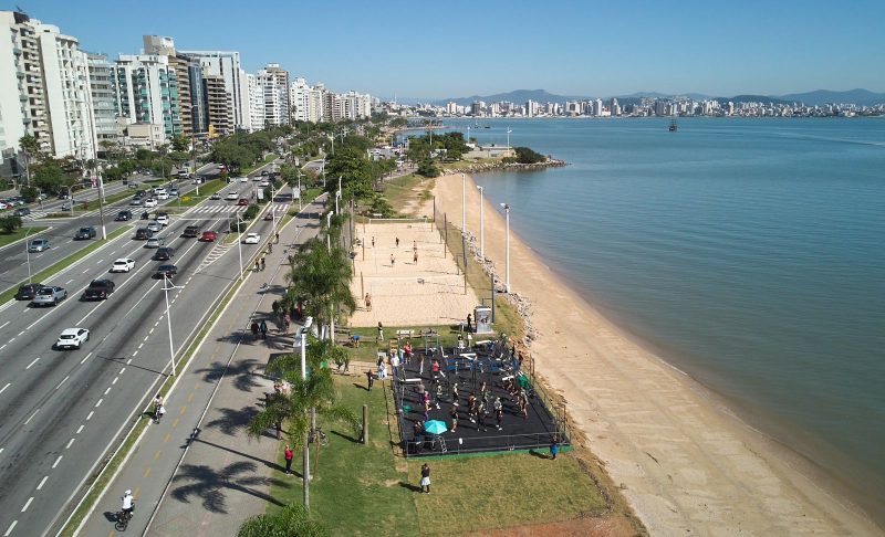 Foto da Beira-Mar Norte, em Florianópolis, de uma perspectiva de cima. No canto direito, aparece o mar, uma pequena faixa de areia e uma área gramada. Ao centro, uma via movimentada com carros. Do lado esquerdo, prédios altos. 