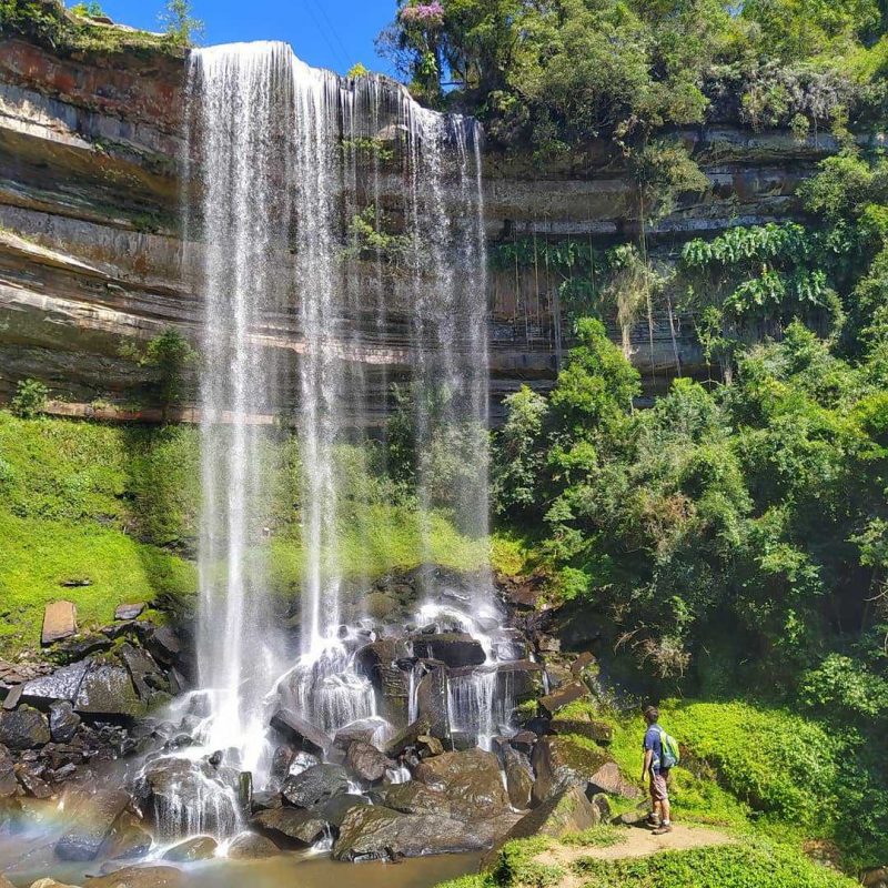 Foto da Cachoeira Paulista, em Doutor Pedrinho. A queda d'água é larga e cai sobre um paredão rochoso laminado. Ao redor há árvores e vegetação. A água cai em algumas rochas e um homem de costas para a foto observa a cachoeira. 