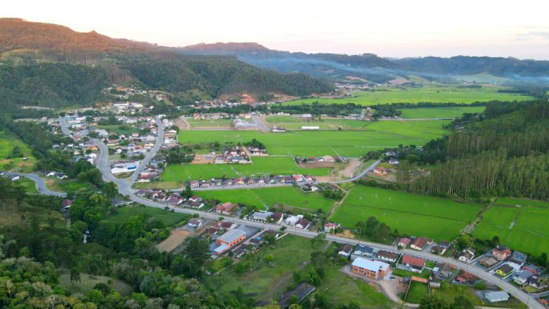 Foto da área urbana central de Doutor Pedrinho, no Vale do Itajaí. Algumas casas e ruas são cercadas por árvores, campos verdes e morros. 