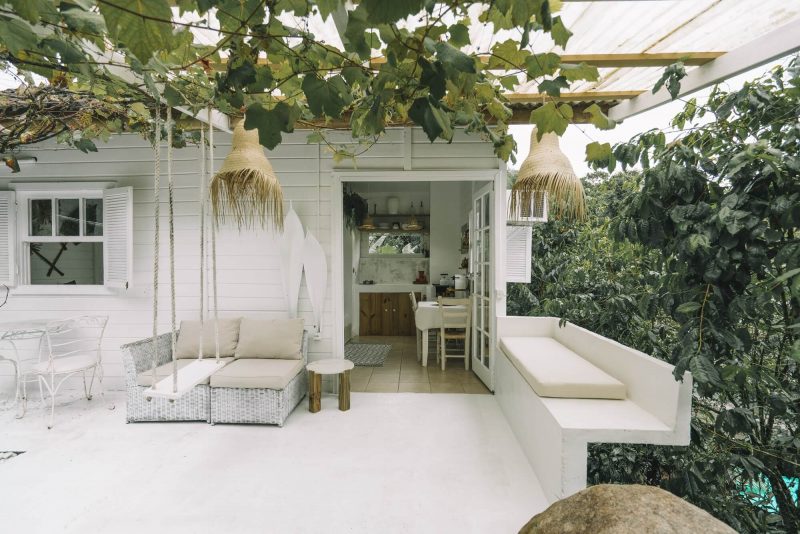 Foto da varanda de um chalé romântico em Florianópolis. Aparece o chalé ao fundo, em madeira e pintado de branco. Na frente dele, uma mesa com duas cadeiras de ferro, um sofá de dois lugares e um banco pendurado no telhado. Tudo é branco, com exceção de plantas trepadeiras verdes espalhadas nas ripas abaixo das telhas do telhado. 