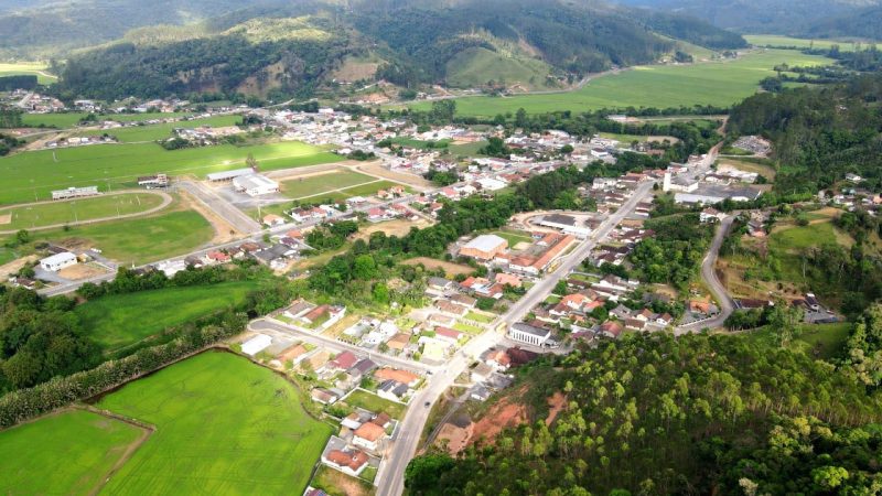 Foto aérea da cidade de Doutor Pedrinho, no Vale do Itajaí. Algumas ruas e casas aparecem entre terrenos gramados e morros verdes 