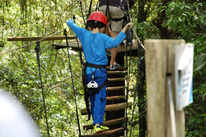 Foto mostra uma criança de costas praticando arvorismo. Ela anda por uma passarela suspensa feita de troncos em meio às árvores no Parque Itá Ecoturismo. O menino veste blusa e calça azuis e um capacete vermelho. Ele se apoia em cordas laterais. 