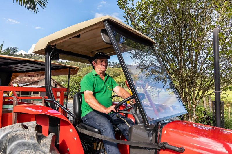 Foto de um trator vermelho sendo dirigido por um senhor branco, de boné preto, camiseta verde e calça jeans. Ao fundo aparece a paisagem de morros verdes e árvores da área rural de Joinville. O sítio oferece passeios com crianças. 