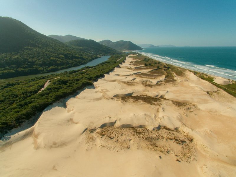 Foto da Praia do Siriú, em Garopaba. No meio da foto, dunas brancas de areia. Do lado direito, o mar. Do lado esquerdo, uma vegetação divide as dunas de areia com uma lagoa estreita e comprida. Após a lagoa e também ao fundo, morros verdes. 