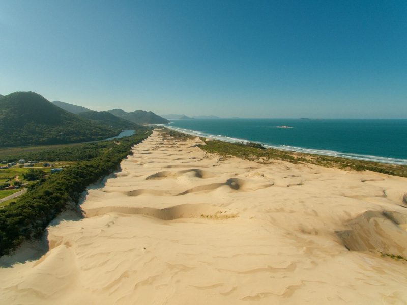 Foto da Praia do Siriú em Garopaba. No centro as dunas de areia. Do lado direito o mar e do lado esquerdo a vegetação e morros verdes, que também aparecem ao fundo.