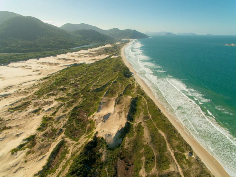 Foto da Praia do Siriú em Garopaba. No meio a extensa faixa de areia com vegetação de restinga. Do lado direito o mar e do lado esquerdo e ao fundo morros verdes. 