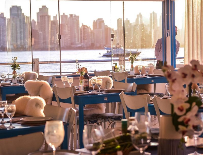 Foto do salão interno do restaurante em Balneário Camboriú. Em primeiro plano aparecem as mesas azuis com taças, pratos e arranjos de flores e cadeiras brancas. Em segundo plano, grandes janelas de vidro deixam ver o mar e os altos prédios de Balneário Camboriú. 