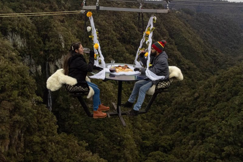 Foto mostra casal bebendo uma taça de vinho a 150 metros de altura, no meio de um vale montanhosa e verde. Eles estão sentados em uma cadeira anexa a uma mesa. Toda a estrutura é presa por cabos de aço.