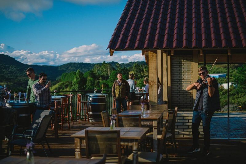 Foto de uma das vinícolas em Santa Catarina, a Monte Agudo - em São Joaquim. A imagem mostra parte de um deck de madeira, com mesas e cadeiras onde um músico toca um violino em pé. Algumas pessoas assistem e fotografam. Ao fundo aparece morros verdes. 